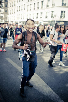 A person wearing a mask and a partially open jacket, revealing their torso, walks towards the camera during the Techno Parade, Edition 2017. People in the background appear to be part of a lively crowd, with buildings and trees visible in the background.