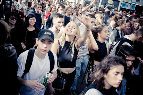 A large group of people are gathered outdoors, many smiling and engaged in conversation. A young woman in the center is wearing a black crop top and making a peace sign with her fingers. The crowd, lively and diverse, as expected at the Edition 2017 Techno Parade, has some holding drinks and backpacks.