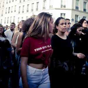 A crowded outdoor scene with people, mostly young women, marching or gathered together in what seems to be a lively 2017 parade. A woman in the foreground wearing a red T-shirt with white text and white shorts stands out. Another woman in black is beside her. The background shows a building facade pulsating with techno beats.
