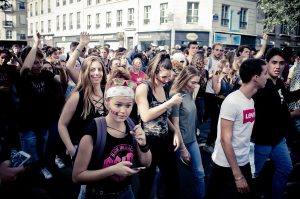 A large group of people is gathered in the street, many looking at their phones. Some individuals are dancing or have their arms raised. The setting appears to be an urban area with buildings and shop signs in the background. The mood seems lively and social, reminiscent of the Edition 2017 Techno Parade.