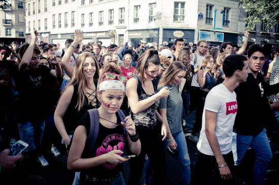 A large group of people is gathered in the street, many looking at their phones. Some individuals are dancing or have their arms raised. The setting appears to be an urban area with buildings and shop signs in the background. The mood seems lively and social, reminiscent of the Edition 2017 Techno Parade.
