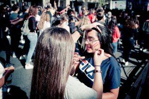 At the Techno Parade, Edition 2017, two women participate in an outdoor event; one is painting red lines on the other's cheek while surrounded by a lively and energetic crowd. The background is filled with people dressed in casual clothing, contributing to the spirited atmosphere.