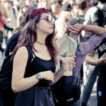A woman with long hair wearing a floral headband, sunglasses, and a black top holds a plastic cup while standing in the crowd at Techno Parade Edition 2017. People in the background are in casual attire, with some facing different directions. The scene appears to be an outdoor event.