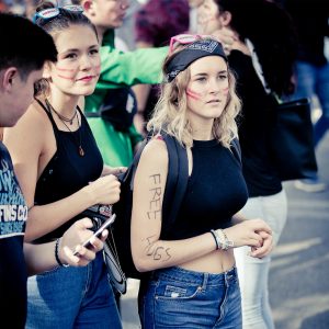 Two women with red face paint and casual black tops participate in the 2017 Techno Parade. One has "FREE HUGS" written on her arm. Both have accessories in their hair, and the background shows a crowd of people socializing.