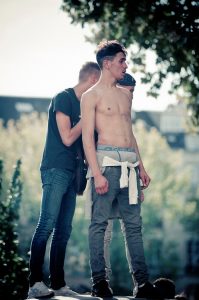 Three young men are outdoors. A shirtless man in the foreground wears jeans with a sweatshirt tied around his waist, reminiscent of a scene from Techno Parade Edition 2017. Behind him, another man in a dark t-shirt and jeans looks down at his phone, while a third person is partially obscured. Trees fill the background.