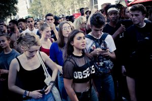 A large group of young people gathered outdoors, featuring diverse individuals. Some are looking ahead while others are engaged with their phones. The central person, wearing a mesh top with "OBSCURITY" written on it, stands out at the Edition 2017 Techno Parade. Trees and buildings provide the backdrop.