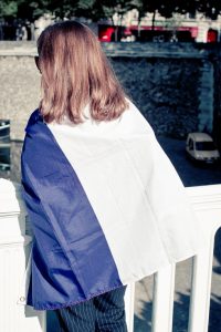 A person with long hair, wearing sunglasses, stands on a bridge at the Techno Parade, facing away from the camera. Draped in a large blue, white, and purple flag from Edition 2017, they create a striking contrast against the stone wall and parked cars in the background.