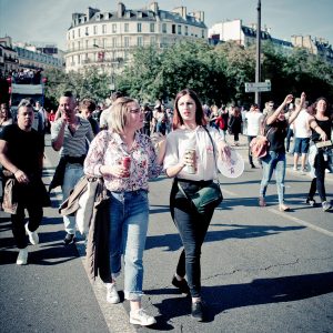 Two women walk and chat in the foreground of a bustling street filled with people attending the Techno Parade, Edition 2017. One holds a drink and wears a patterned blouse, while the other carries a can and wears a black shirt. Buildings and trees can be seen in the background on a sunny day.
