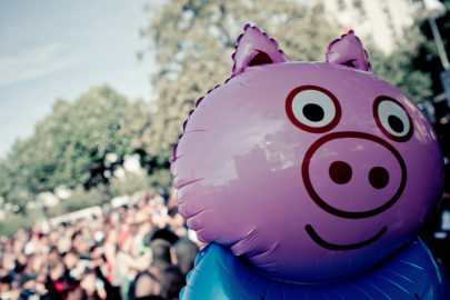 A large, pink inflatable pig with cartoonish eyes and a smiling face is displayed in the foreground. In the background, an out-of-focus crowd of people can be seen enjoying the Techno Parade, Edition 2017, alongside some trees under a partly cloudy sky.