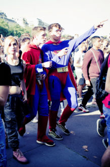 A crowd of people, including two wearing superhero costumes and capes, are walking outside. One person in a blue and red superhero outfit points to something in the distance, while others in casual clothes walk alongside them. It's the Techno Parade: Edition 2017, with trees and buildings in the background.