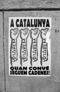 A black and white poster on a wall with the text "A CATALUNYA VOTAREM VOTAREM VOTAREM VOTAREM QUAN CONVÉ SEGUEM CADENES!" above four raised fists, symbolizing the Référendum de 2017 for the indépendance de la Catalogne.