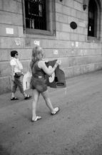 A black-and-white photo of two people walking along a sidewalk beside a stone building in Barcelone. The person in front is carrying a large, partially folded cardboard cutout of a bottle. Both are dressed casually, and there are posters and graffiti on the building's wall, possibly referencing the indépendance de la Catalogne or the Référendum de 2017.