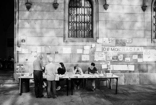 Black-and-white photo of people sitting at a long table with paperwork in front of a stone building. The wall behind them is covered with various signs and papers, including a large sign reading "DEMORACIA!" The setting suggests an outdoor public event in Barcelone, likely related to the Référendum de 2017 sur l'indépendance de la Catalogne.