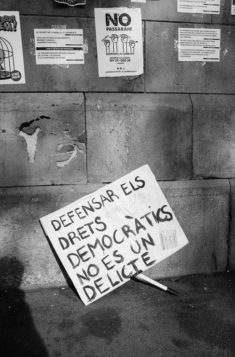 A black and white photo shows a protest sign leaning against a street wall plastered with posters in Barcelone. The sign in Catalan reads, "Defensar els drets democràtics no és un delicte," echoing the sentiments from the Référendum de 2017 for the indépendance de la Catalogne.
