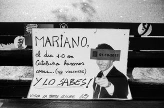 A black and white photo of a protest sign on a bench. The sign features a photo of a man pointing forward with the text in Spanish, "Mariano, el día 1-O en Cataluña haremos cosas...(no violentas) Y LO SABES! Visca la terra lliure". A date "01-10-2017" is also mentioned, referencing the Référendum