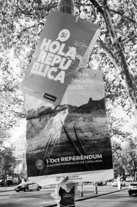 Black-and-white image of two posters on a lamppost. The top poster says "Sí HOLA REPÚBLICA" and promotes catalunyasi.cat. The bottom poster advertises the 1st October référendum de 2017 for Catalan self-determination, depicting train tracks leading into the distance.