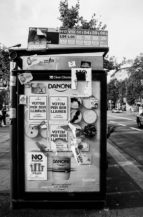 A bus stop advertising board in a city, covered with numerous posters and stickers. Some posters include political messages advocating for the Référendum d'indépendance in Catalogne and freedom, while others feature product advertisements. The surrounding area includes a street and trees.