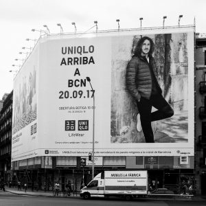 Black and white photo of a grande affiche in a city. The large Uniqlo billboard announces Uniqlo's arrival in Barcelona on September 20, 2017, featuring a man in a winter jacket. Below the billboard, there is a street and a mobile furniture store advertisement.