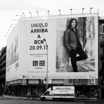 Black and white photo of a grande affiche in a city. The large Uniqlo billboard announces Uniqlo's arrival in Barcelona on September 20, 2017, featuring a man in a winter jacket. Below the billboard, there is a street and a mobile furniture store advertisement.