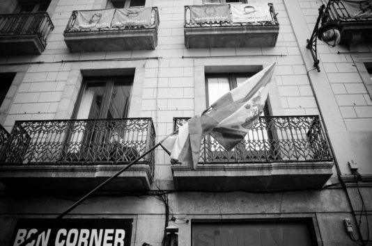 A black and white photo from 2017 depicts a flagpole leaning diagonally across the front of a building with ornate iron balconies in Catalogne. Two closed windows are visible on the second floor, with banners partially draped over the railings, hinting at the référendum d'indépendance. The lower part features signs and windows.