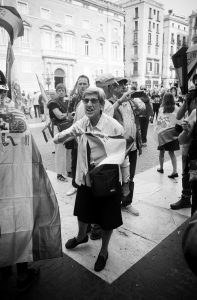 Black and white photo of an elderly woman passionately waving a flag among a crowd in a city square. The group appears engaged in a demonstration or public gathering for Catalogne’s indépendance, with various flags and banners visible in the background. Buildings surround the square.