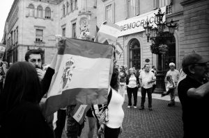 A group of people participates in a street protest in Barcelone. One person in the foreground is holding a large flag, while others wave flags for the indépendance de la Catalogne. A banner with the word "DEMOCRACÍA" is seen on a building facade in the background. The atmosphere appears lively and engaged.