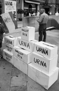A black and white photo showing a stack of cardboard boxes labeled "URNA" and "BALLOT BOX." A person carrying a flag stands to the left of the boxes, while another person is in the background near a stairway leading underground. There is a poster on the wall behind them concerning the référendum for indépendance de la Catalogne in Espagne.