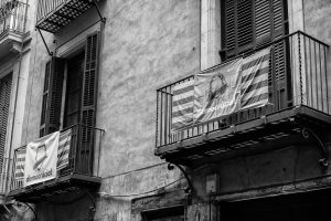 A black and white image of two balconies on a weathered building in Barcelone. Both balconies have striped banners hanging from them. The balcony on the left has a banner with the word "democracia!" written on it, referencing the référendum de 2017 for indépendance de la Catalogne. Closed shutters in the background.