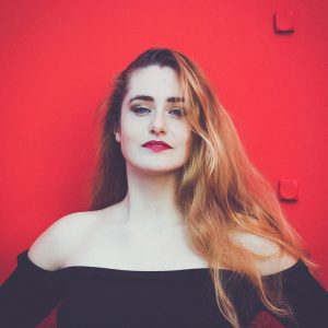 A woman with long, wavy hair and bold makeup stands against a bright red background reminiscent of Calder's vibrant aesthetics. She wears a black, off-the-shoulder top and gazes confidently at the camera, embodying the essence of Clotilde.
