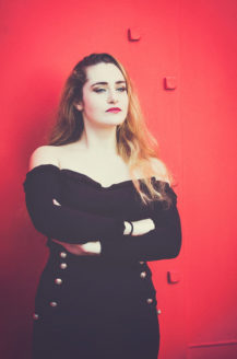 A woman with long, wavy blonde hair, reminiscent of Clotilde, stands confidently against a vibrant red wall. She is wearing an off-the-shoulder black top and has her arms crossed. Her expression is serious and focused, evoking the dramatic presence of Calder's araignée rouge.