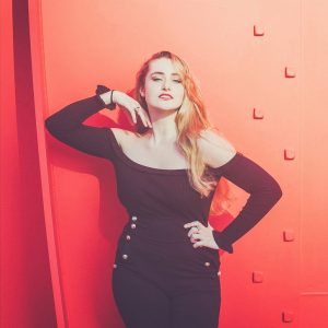 A woman with long blonde hair poses confidently against a vibrant red background, reminiscent of Calder's bold art style. She is wearing a black off-the-shoulder top and high-waisted black pants adorned with buttons. Her right arm is bent, hand resting near her neck, while her left hand rests on her hip.