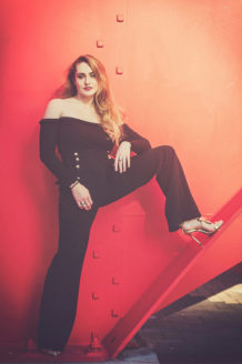 A woman with long hair poses confidently against a red, industrial background reminiscent of Calder's "Araignée rouge." She is dressed in a stylish, black off-the-shoulder jumpsuit with button details and wearing high heels. One leg is bent, resting on an angled beam, with her hand on her thigh, evoking the elegance of Clotilde.