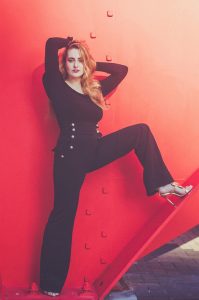 A woman with long, wavy hair poses confidently against a large red metal structure reminiscent of Claudel's "araignée rouge." Clotilde is dressed in a stylish black outfit with metallic button accents and wears high-heeled sandals. One leg is raised, resting on part of the structure, and her arms are positioned overhead.