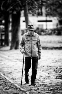 A black-and-white photo by Robert Hold captures an elderly person walking on a leaf-strewn path. They are wearing a jacket, a hat, and are using a walking stick. Trees and blurred background elements suggest a park or outdoor setting.