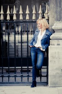 A woman with blonde hair, wearing a blue leather jacket, white blouse, and denim jeans, stands casually against a pillar next to an iron fence. She has her hands in her pockets and is looking to her right, smiling. The background features a building entrance.