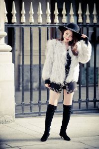 A woman wearing a fur coat, black wide-brimmed chapeau, and black knee-high boots stands in front of a wrought-iron fence. She is holding the brim of her hat with one hand and slightly smiling. The background includes a stone pillar and some architectural details.