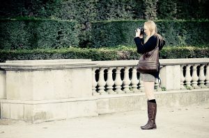 A woman with long blonde hair, dressed in a dark coat, gray skirt, and brown knee-high boots, stands near a balustrade labeled "Sans Titre," holding a camera up to her face and taking a photo. There is a hedge in the background.