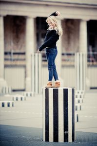 Lily-Rose, a young girl with long blonde hair, stands balanced on a black-and-white striped pedestal in an urban area, her arm raised gracefully. Dressed in a black sweater, blue jeans, and brown boots, she s'amuse against the backdrop of more pedestals and a classical building.