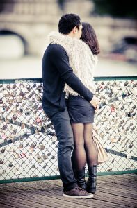 Pierre et Anna stand close together on a bridge, holding each other and looking out over the water. The bridge's railings are adorned with numerous love locks. Pierre is dressed in dark clothing, and Anna wears a fur jacket, black skirt, and tights.