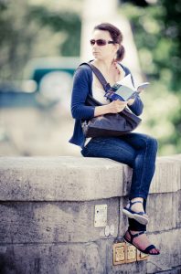 A woman in sunglasses sits on a stone ledge, holding an open book. She wears a blue cardigan, white shirt, jeans, and sandals. Her crossbody bag lies across her shoulder. She gazes to her right, with a blurred, green outdoor background behind her. This scene captures the serene style of Inès Diaz.