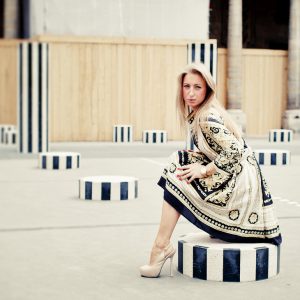 A woman with long blonde hair, wearing a beige and black patterned robe and high heels, sits on a black and white striped cylindrical structure in an outdoor urban setting. More similar striped cylinders are scattered around her.