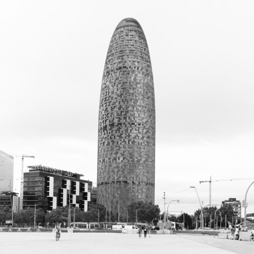 Black and white image of the tour Agbar, a tall, cylindrical building with a curved top and a mosaic-like exterior in an urban area of Barcelone. Bicyclists and pedestrians are visible in the foreground, with surrounding modern buildings and construction cranes in the background.