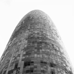 Black and white image of a tall, modern, cylindrical building with a rounded top. The facade consists of many small, reflective windows creating a pattern across the structure. The perspective is looking up from the base of the iconic Tour Agbar in Barcelone, Espagne.