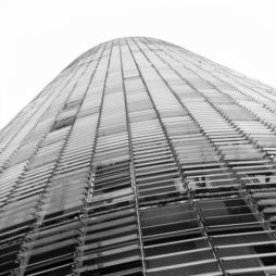A low-angle view of a modern, high-rise building in Barcelone with a sleek, curved facade. The structure features horizontal lines and a grid-like pattern, reminiscent of the iconic tour Agbar, creating an impression of futuristic architecture against a stark white sky in Espagne.