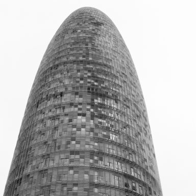 A black-and-white photograph of the sleek, bullet-shaped tour Agbar in Barcelone, with its distinctive grid-like exterior surface resembling overlapping tiles or panels. The building narrows towards the top, giving it a modern appearance against a white sky.