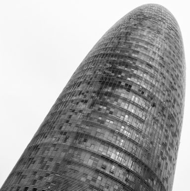 A tall, cylindrical, modern skyscraper with a curved, tapering top, covered in a grid-like pattern of windows and reflective surfaces, set against an overcast, grey sky. The building’s sleek design creates a futuristic and imposing appearance reminiscent of the Tour Agbar in Barcelone, Espagne.