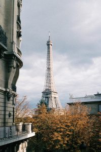 A view of the Eiffel Tower on a cloudy day with autumn-colored trees in the foreground and classic Parisian buildings on either side, capturing the quintessential charm of Paris.