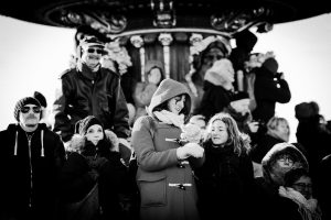 A group of people bundled in winter clothing stand closely together outdoors, some smiling and interacting, with a carousel or ornate structure in the background. The image is in black and white.