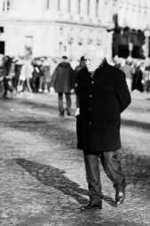 An older man wearing a dark coat and trousers walks alone on a cobblestone street, hands in his pockets. In the background, a crowd of people stands near a large building in bright sunlight. The photo is in black and white.