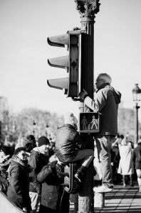 A man and a child climb a traffic light pole while a crowd gathers below on a city street. The scene appears to be during a public event or protest. The image is in black and white.